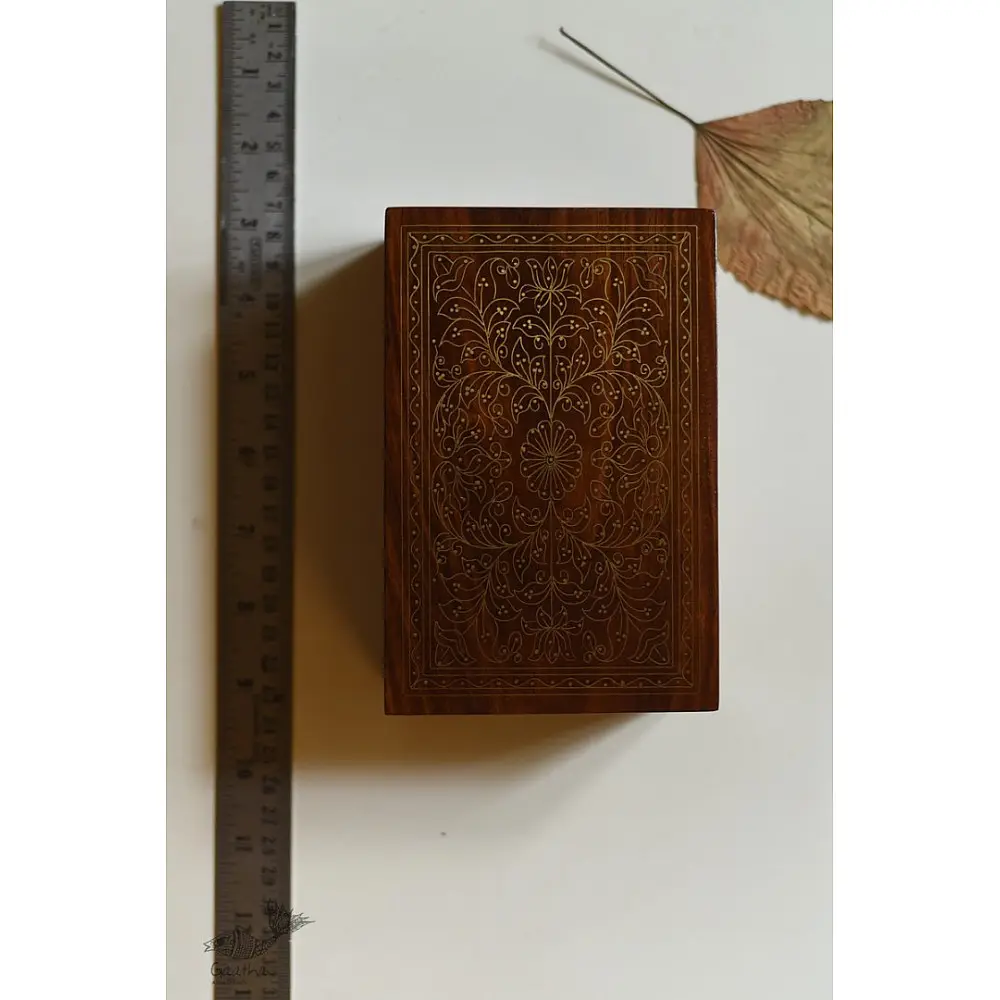 handcrafted rectangular wooden box featuring complex brass wire Tarkashi inlay work forming a central paisley floral layout and bordering coin circles next to a dry leaf