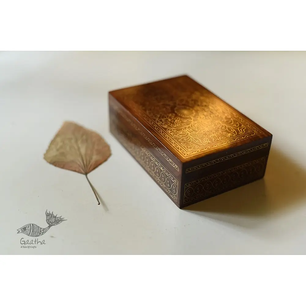 handcrafted rectangular wooden box featuring complex brass wire Tarkashi inlay work forming a central paisley floral layout and bordering coin circles next to a dry leaf