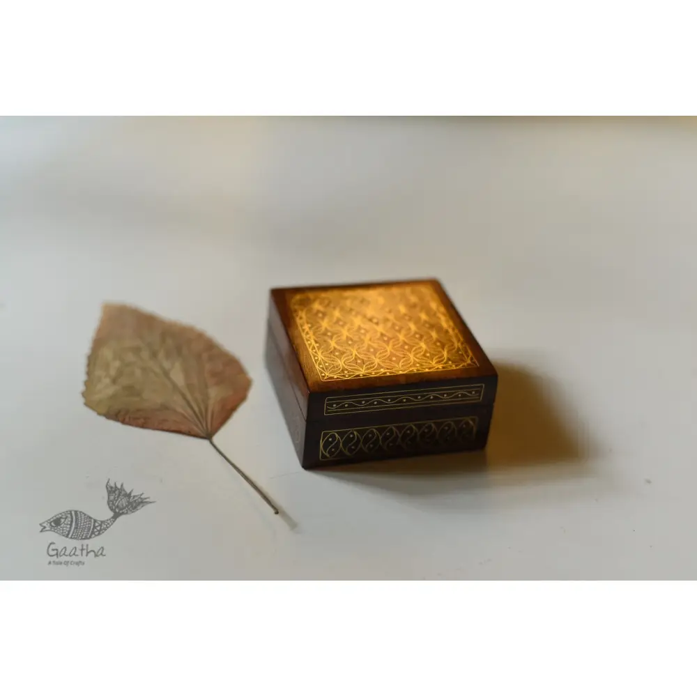 handcrafted square wood gift box with intricate geometric brass wire Tarkashi inlay work forming a repeating overlapping circle pattern across the polished lid next to a dry leaf