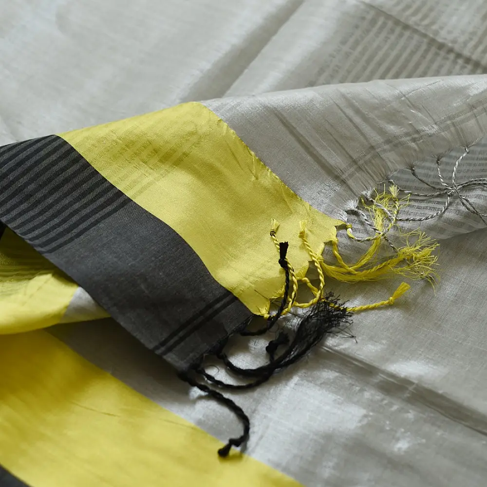 handloom pure cotton saree in soft grey and white featuring a bright lemon yellow color blocked panel with a black striped zari border and handcrafted tassels