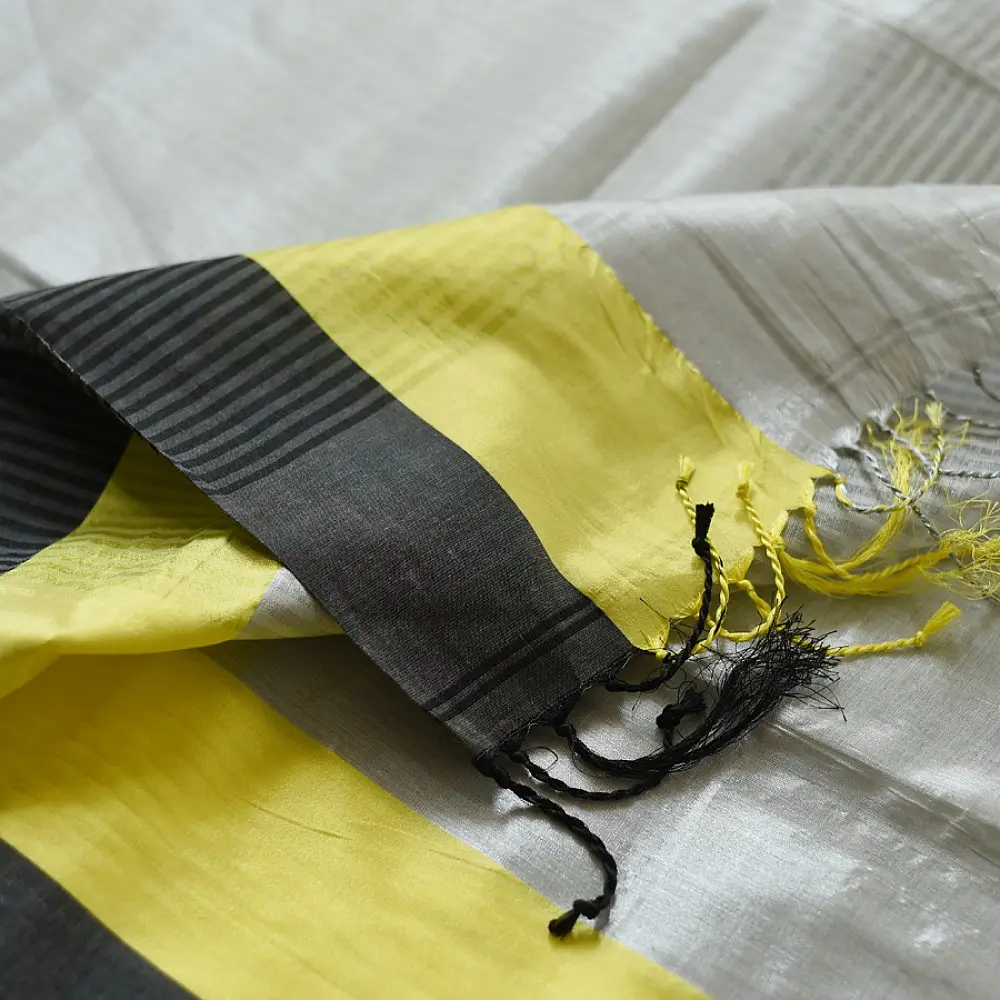handloom pure cotton saree in soft grey and white featuring a bright lemon yellow color blocked panel with a black striped zari border and handcrafted tassels