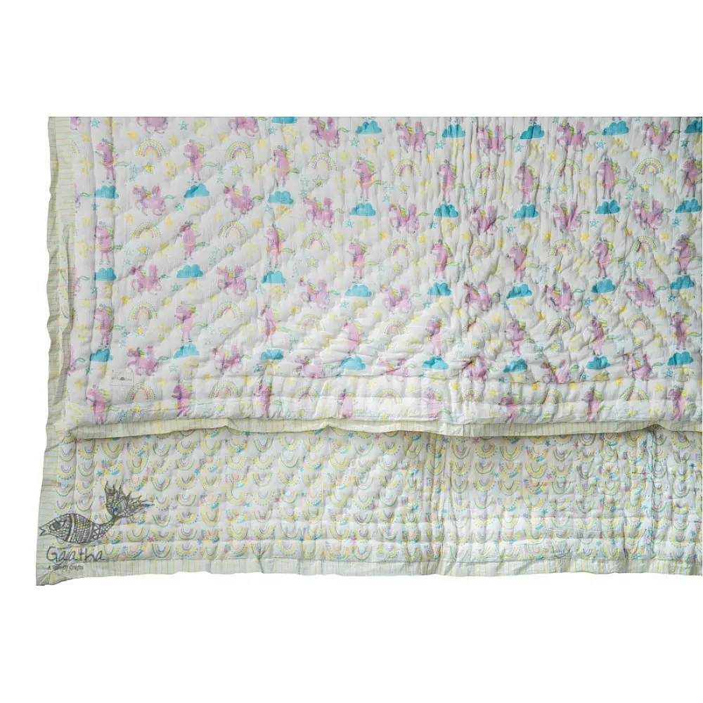 mauve and white block printed baby quilt with whimsical unicorns and rainbows pattern draped over a crib
