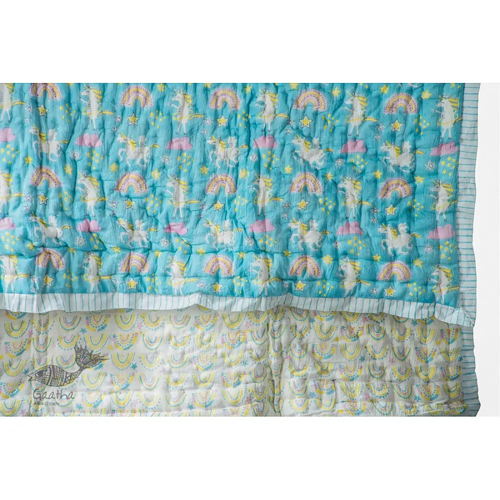 blue and teal block printed baby quilt with whimsical unicorns and rainbows pattern draped over a crib