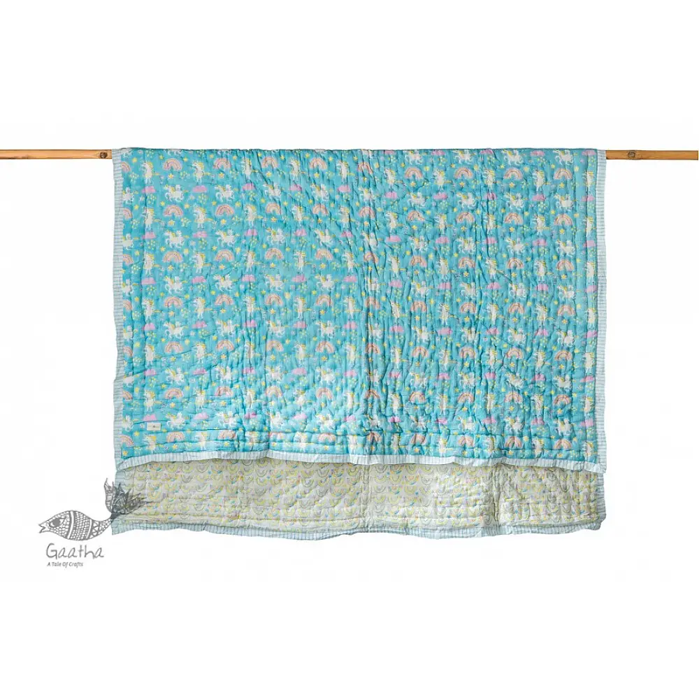 blue and teal block printed baby quilt with whimsical unicorns and rainbows pattern draped over a crib