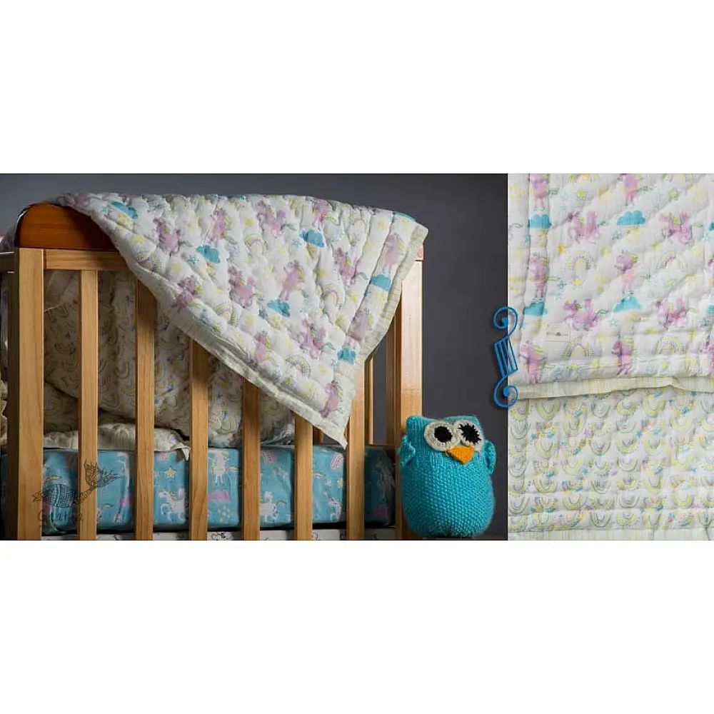 mauve and white block printed baby quilt with whimsical unicorns and rainbows pattern draped over a crib